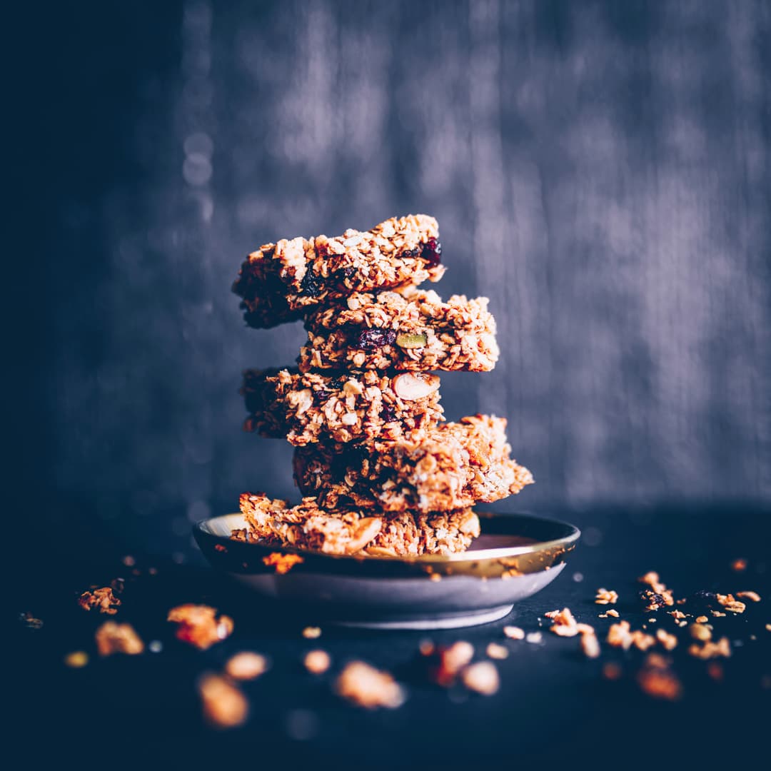 Stack of homemade granola bars with oats, nuts, and dried fruits on a small plate, with scattered crumbs on a dark background